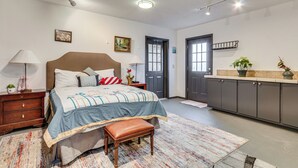 Une chambre à coucher comprenant un lit, des tables de chevet, un banc, une commode, une lampe, un tableau, une porte, une fenêtre et un coin cuisine avec des placards et un plan de travail.