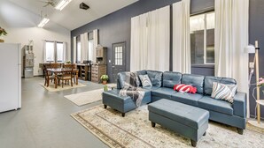 Un salon moderne avec un canapé d’angle, un pouf, une table à manger avec des chaises, et un coin cuisine en arrière-plan.