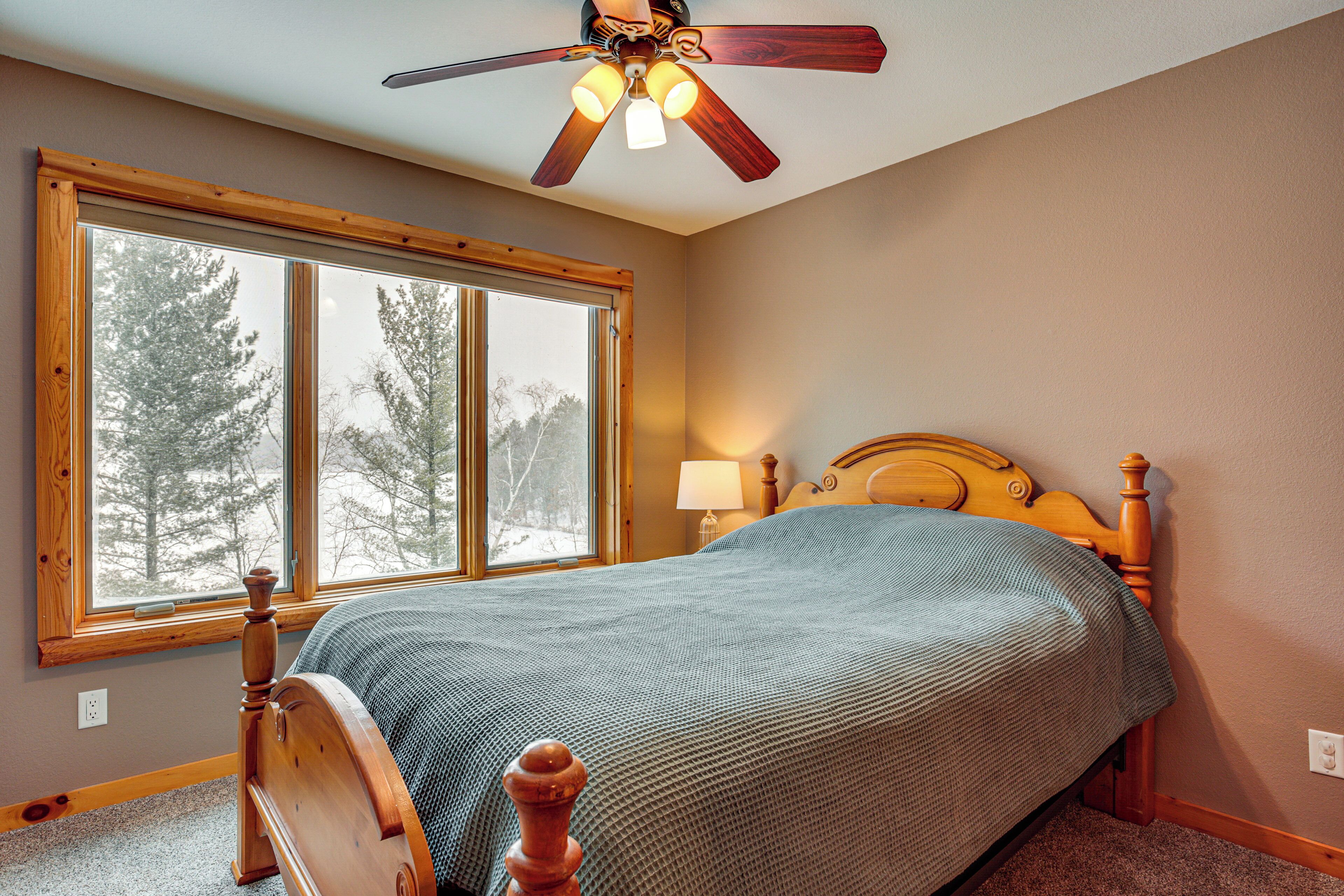 A bedroom with a wooden bed, a ceiling fan, and a large window with a view of trees.