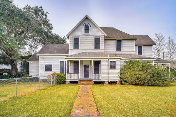 A two-story house with a front porch, a brick pathway leading to the entrance, and a fenced yard. - Historic Home w/ Wraparound Porch + Private Yard! (Jeanerette)