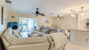 A living room with a beige sofa, a coffee table, and a TV. There are ceiling fans, a kitchen area with cabinets, and a door leading to another room.