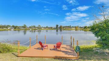 A wooden dock with two red Adirondack chairs facing a calm lake, surrounded by greenery and a clear blue sky.
