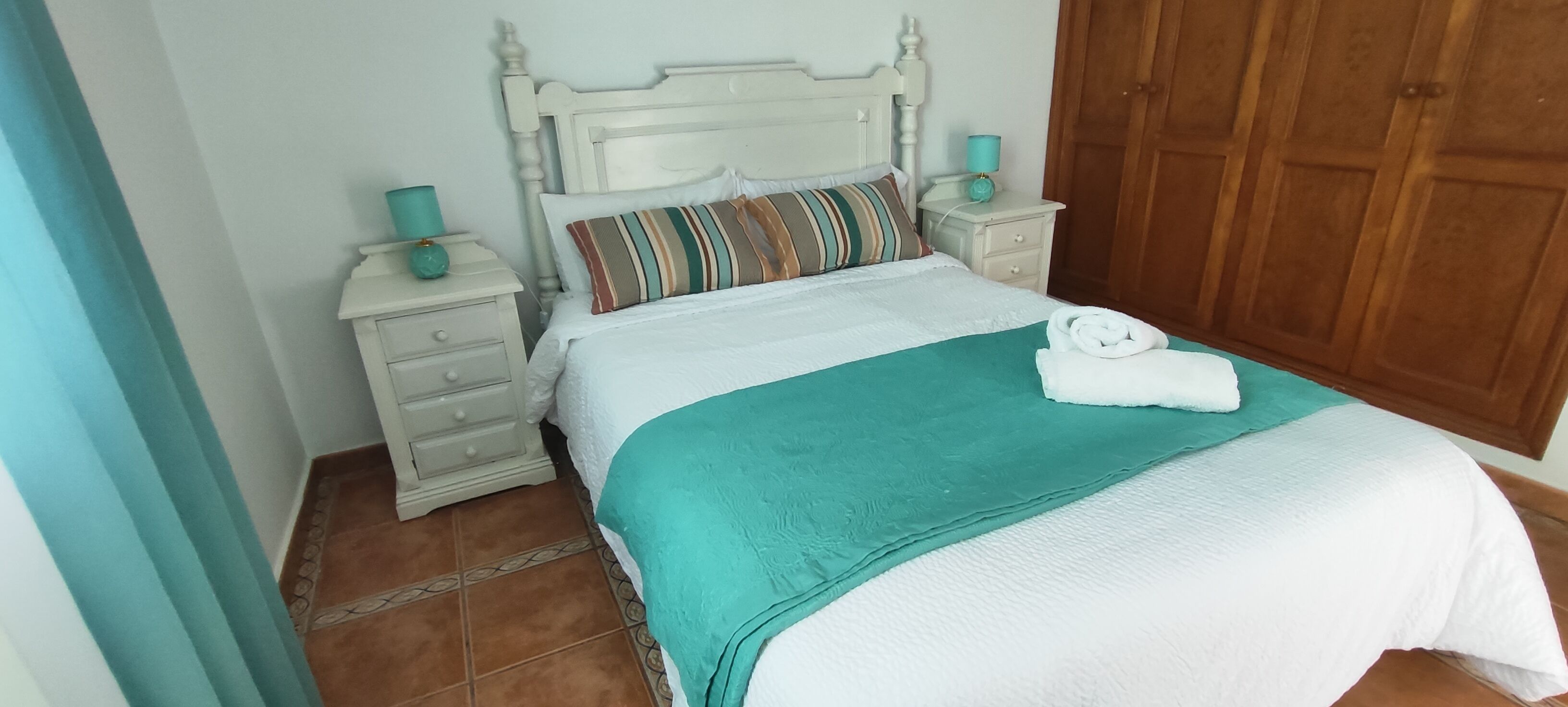 Ein ordentlich bezogenes Bett mit einem gestreiften Kopfkissen, einer türkisfarbenen Decke und einem gefalteten Handtuch auf einer weißen Bettdecke. Neben dem Bett steht ein weißer Nachttisch mit einer türkisfarbenen Lampe.