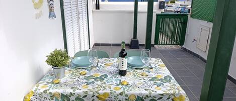 A dining table set with plates, glasses, and a bottle, surrounded by chairs, on a balcony with a view of the sea.