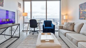 Et moderne hotelværelse med et stort vindue, et fladskærms-tv, en blå lænestol, en beige sofa og et træsofabord.