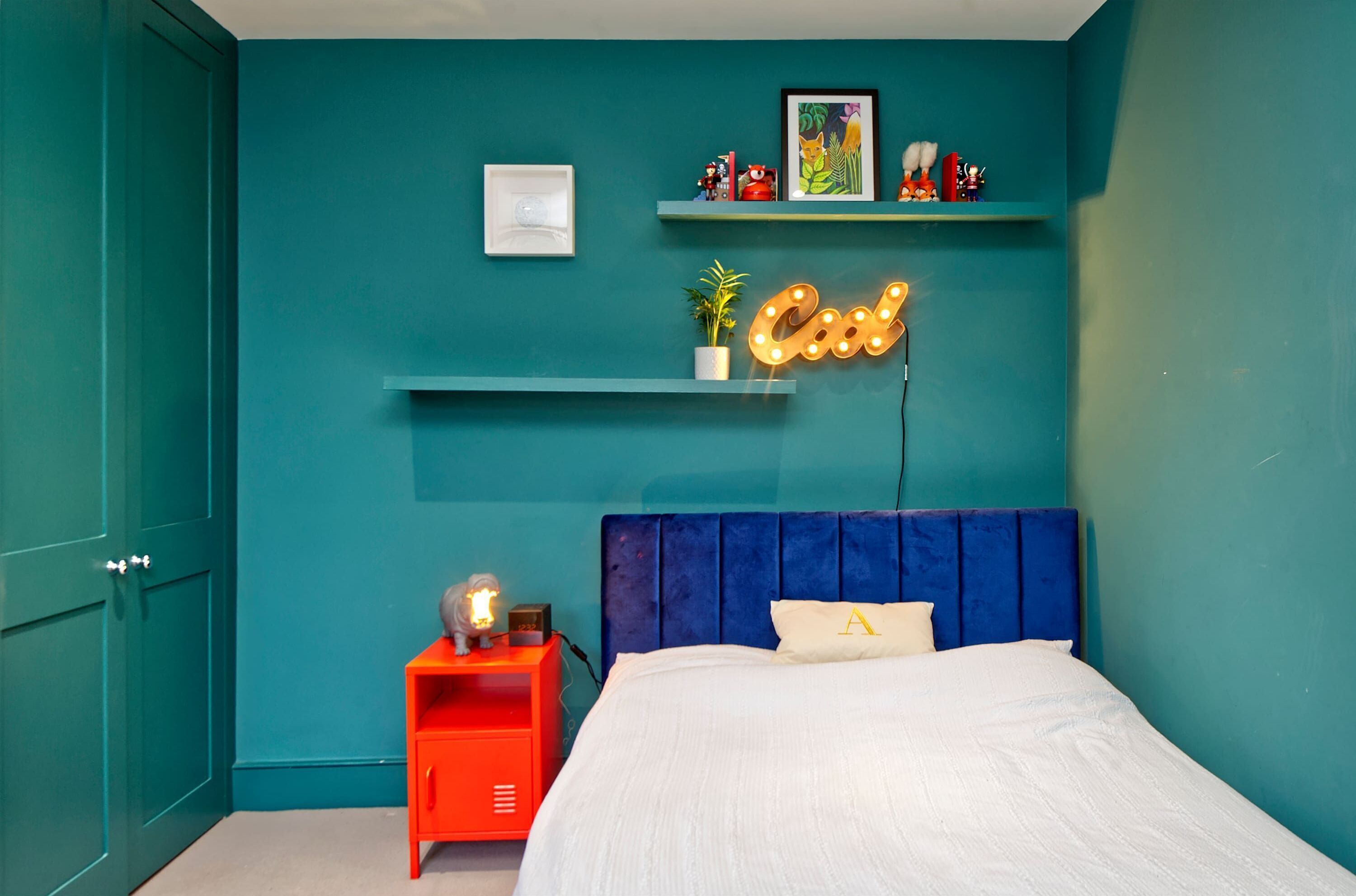Ein Schlafzimmer mit türkisfarbenen Wänden, einem Bett mit blauer Kopfteil, einem Neon-Schild und einem Nachttisch in Rot.