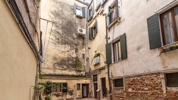 A narrow alleyway with buildings on either side, featuring a mix of brick and plaster walls, some with shutters and potted plants. - Ca Nova II by Wonderful Italy (Venecia)