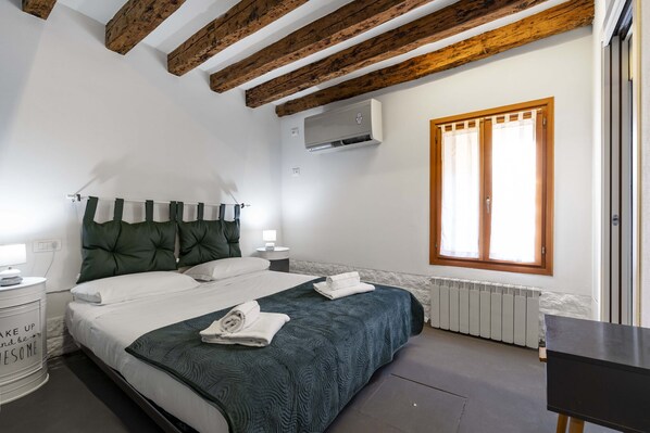 A bedroom with a bed, a nightstand, an air conditioning unit, a window, and a radiator.