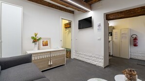 A modern hotel lobby with a grey sofa, a wooden cabinet with a vase of flowers, a flat-screen TV mounted on the wall, and a fire extinguisher. - Ca Nova II by Wonderful Italy (Venecia)