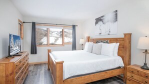 Een slaapkamer met een houten bed, een televisie op een houten dressoir, een nachtkastje met een lamp en een raam met gordijnen.