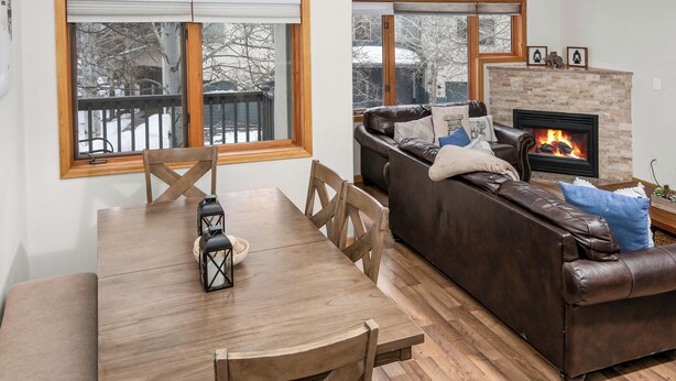 Een gezellige woonkamer met een open haard, een eethoek met een houten tafel en stoelen, en een leren bank met kussens.