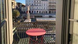 Un balcon avec une table et des chaises rouges, donnant sur une marina où l’on aperçoit des bateaux et des bâtiments en arrière-plan.