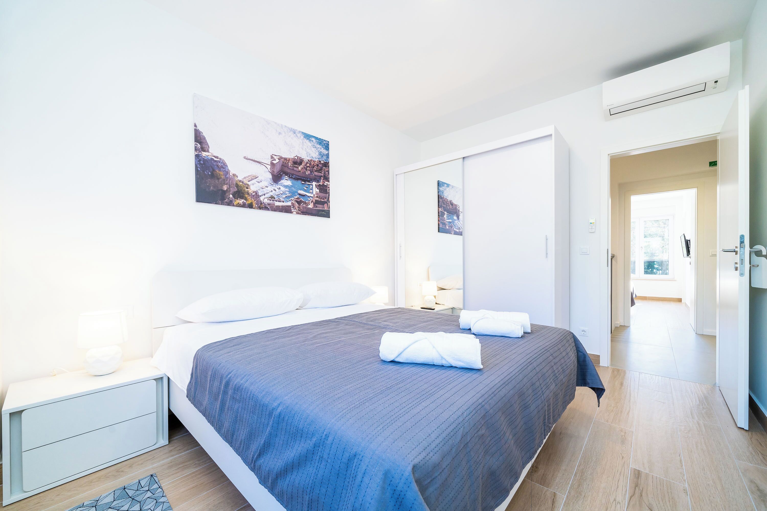 A modern hotel room with a large bed, a bedside table with a lamp, a wall-mounted air conditioner, and a door leading to another room.