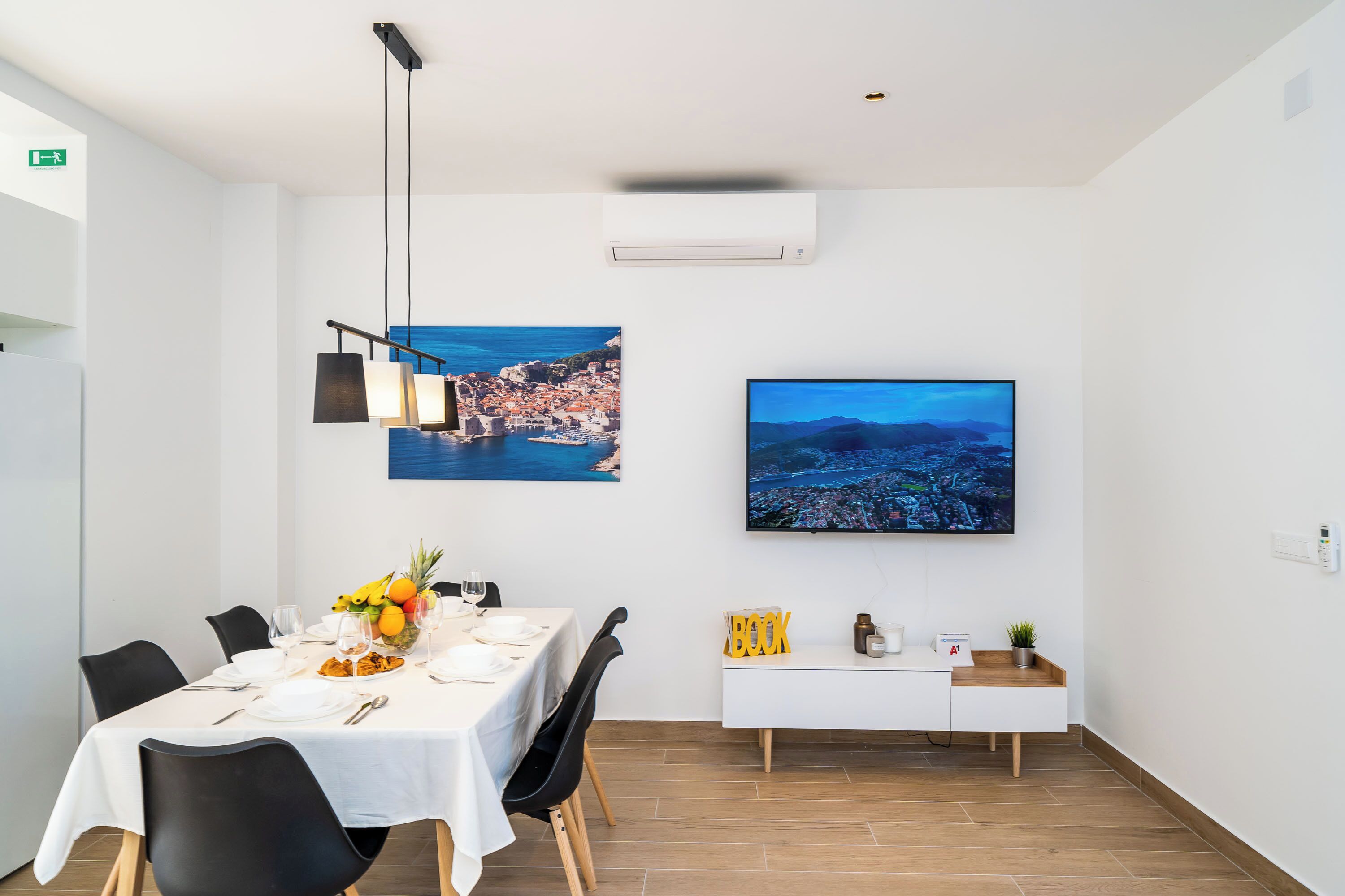 Un comedor con una mesa blanca, sillas negras y un mueble blanco que alberga un televisor y una planta.