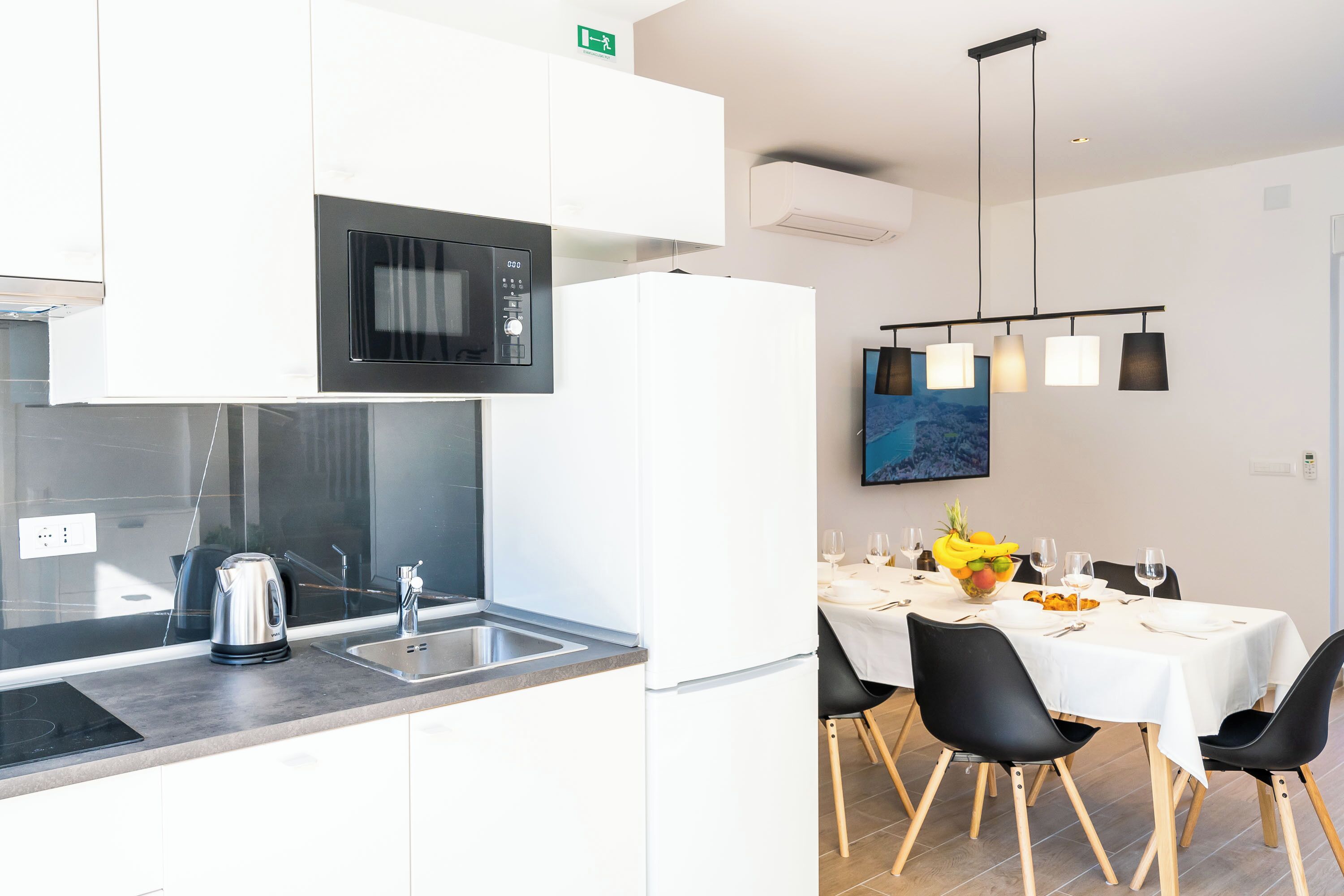 A modern kitchen with a built-in microwave, kettle, and sink. There is a dining area with a table set for two, chairs, and a painting on the wall.