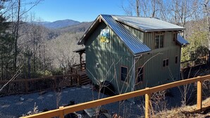 A rustic cabin with a metal roof, surrounded by a wooden deck and gravel ground, set against a backdrop of trees and mountains. - Rocking on Heaven's Door-Covered Porch -  WIFI - Gas Grill (Topton)