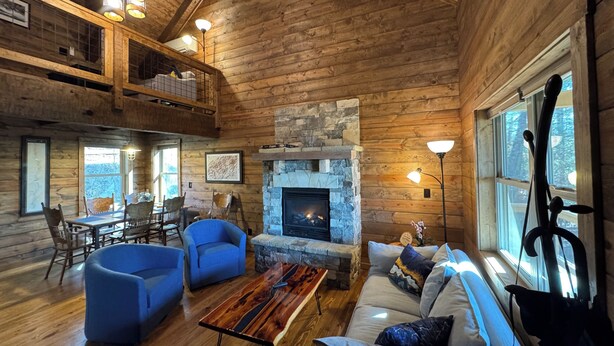 A spacious cabin interior with a stone fireplace, wooden walls, and a dining area with blue chairs. - Rocking on Heaven's Door-Covered Porch -  WIFI - Gas Grill (Topton)
