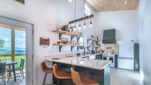 A modern kitchen with a central island, bar stools, wooden shelves, and a view through the glass door. - Brook Haven-Mountain Views - Hot Tub - Outdoor Pizza Oven (Bryson City)