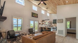 A spacious living area with a large brown leather sofa, a dining table, and chairs. There is a kitchen area with a stove and a refrigerator. The room has large windows and a ceiling fan. - Brook Haven-Mountain Views - Hot Tub - Outdoor Pizza Oven (Bryson City)