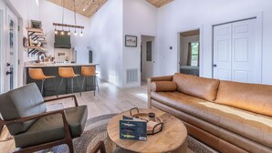 A modern living room with a leather sofa, a wooden coffee table, and a kitchen area with bar stools. - Brook Haven-Mountain Views - Hot Tub - Outdoor Pizza Oven (Bryson City)