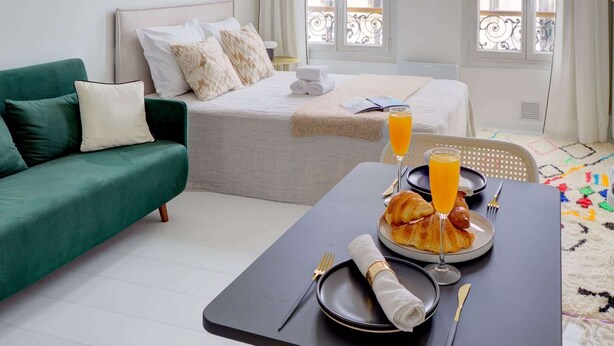 A hotel room with a bed, a green sofa, a dining table with breakfast, and large windows.