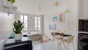 A compact living space with a bed, a dining area with a table and chairs, a washing machine, and a potted plant.