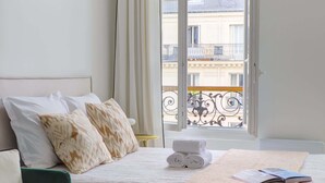 A bedroom with a bed, a green armchair, a window with curtains, and a view of buildings outside.