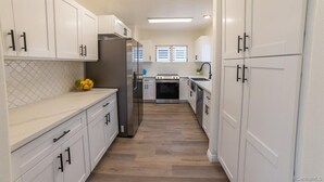 A modern kitchen with white cabinets, a stainless steel refrigerator, a built-in oven, and a window with blinds.