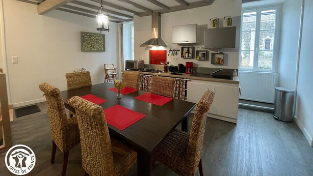 A dining area with a dark wooden table and wicker chairs, a kitchen with modern appliances, and a view of the outside through a window.