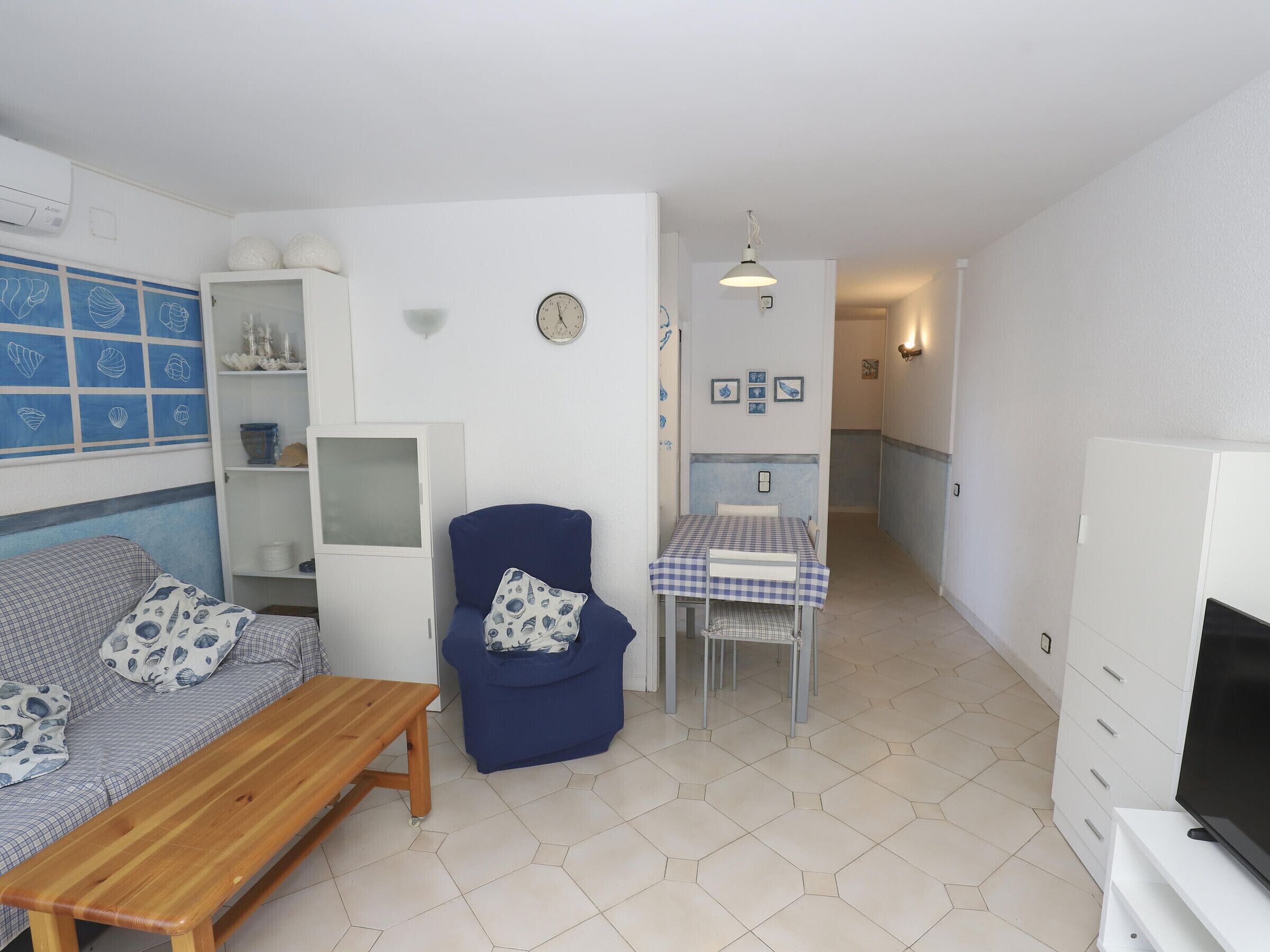 Un espace de vie compact comprenant un canapé, un fauteuil bleu, un banc en bois, une petite table à manger recouverte d’une nappe à carreaux, un téléviseur et une horloge murale.