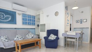 Un salon comprenant un canapé, une table en bois, un fauteuil bleu, une petite table à manger recouverte d’une nappe à carreaux, et un mur orné d’une horloge et de tableaux encadrés.