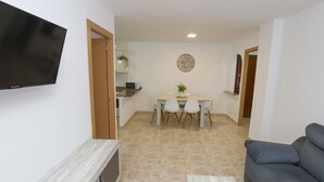 Un salon avec un canapé gris, un téléviseur fixé au mur et un coin repas comprenant une table et des chaises.