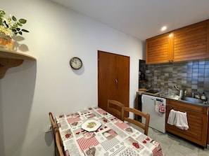 A compact kitchen with wooden cabinets, a small dining table with a checkered tablecloth, and a clock on the wall.