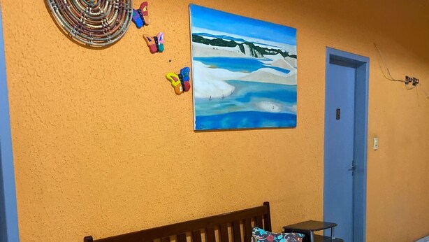 A wooden bench with colorful cushions against an orange wall, a painting of a snowy landscape, and a woven wall hanging. - Pousada santos  (Barreirinhas)