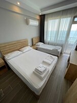 A hotel room with two beds, a wooden headboard, a large window with sheer curtains, and a view of the sea.
