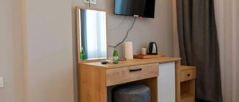 A compact room with a wooden desk, a mirror, a television mounted on the wall, and a small stool.