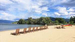 A beach with rows of wooden lounge chairs facing the water, a row of kayaks, and a mountain range in the background. - Kids Love Hidden Bunk Room - Seasons at Sandpoint (Sandpoint)