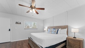 A bedroom with a wooden bed, a nightstand with a lamp, a ceiling fan, a window with blinds, and a door. - Sc11-silver Cove 11 1 Bedroom Condo (Branson)