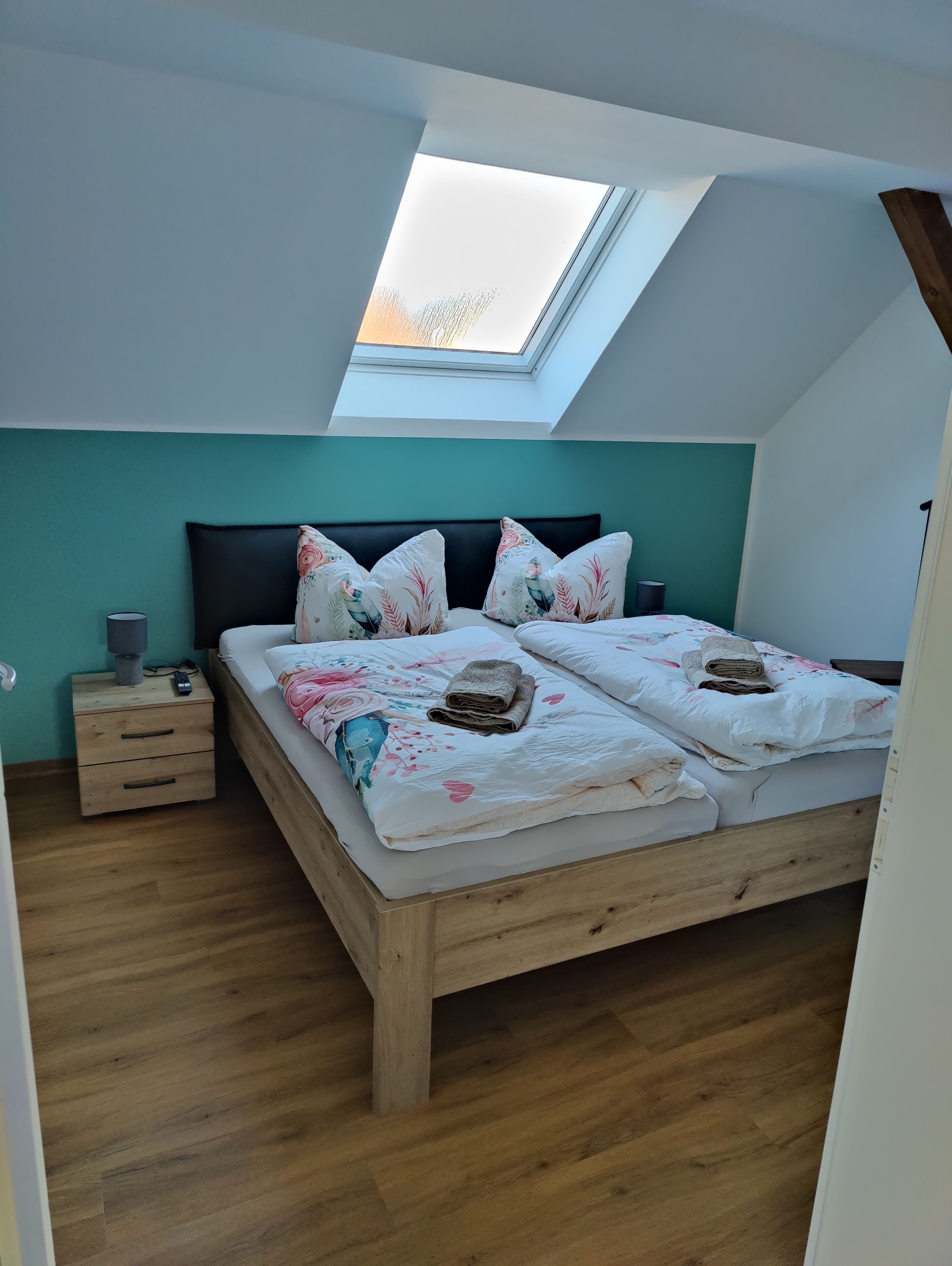 A bedroom with a skylight, a bed with floral bedding, a nightstand, and a wooden floor.