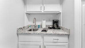 Una cocina compacta con armarios blancos, encimera de granito, pileta de acero inoxidable, cafetera y dispensador de toallas de papel.