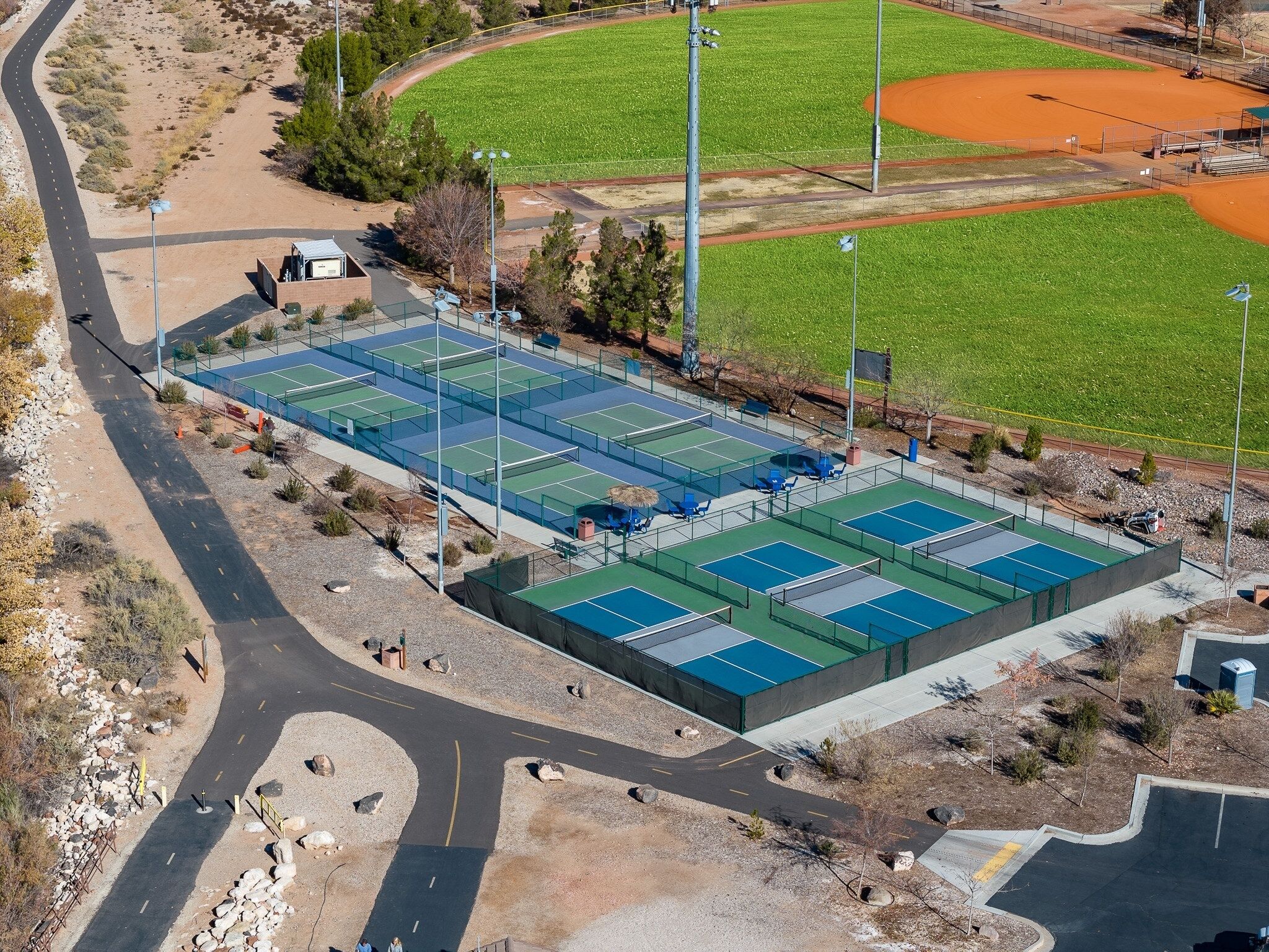 Vista aérea de un complejo deportivo con varias canchas de tenis, un campo de béisbol y una carretera con una curva.