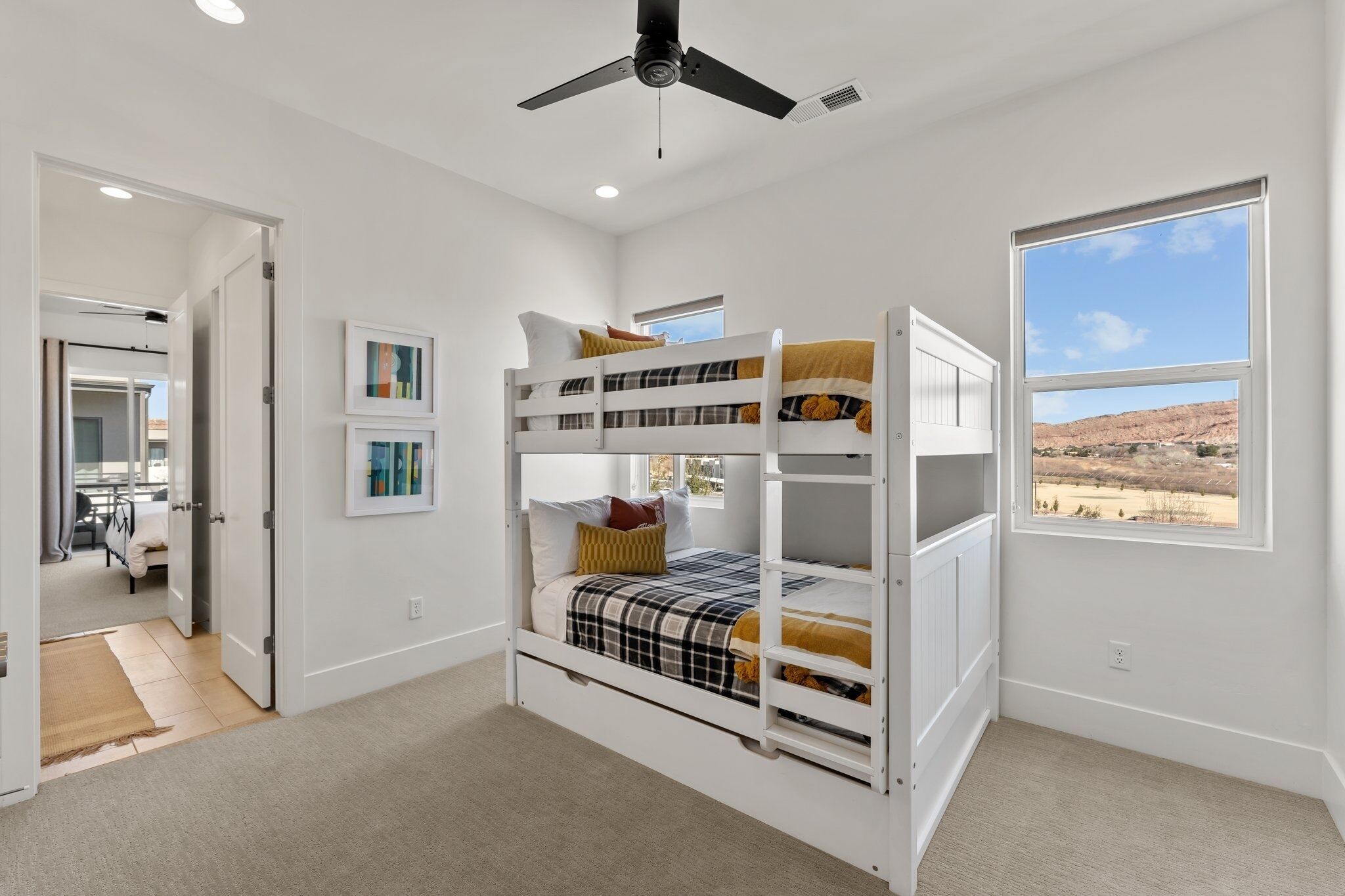 A bunk bed room with a ceiling fan, a window with a view of the desert, and a door leading to another room.