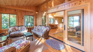 Interior acogedor de una cabaña con paredes y suelos de madera, zona de comedor y grandes ventanales con vistas al bosque.