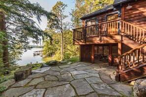 A cabin with a stone-paved deck, a wooden balcony, and a view of a lake surrounded by trees. - Lovely Black Cat Island waterfront (Moultonborough)