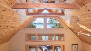 Una habitación con techo inclinado, vigas de madera y vista a un lago a través de un tragaluz.