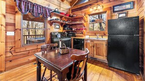Interior de una cabaña de madera con una mesa de comedor, sillas, un refrigerador, una estufa y una ventana que da a un paisaje con árboles.