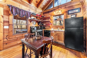Interno di una baita di legno con un tavolo da pranzo, sedie, un frigorifero, un fornello e una finestra vista alberi.