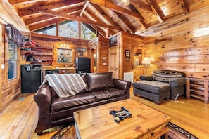 Interno di una baita di legno con un divano in pelle marrone, un tavolino in legno e un camino.