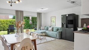A modern living room with a sofa, dining table, and chairs. There is a kitchen area with a refrigerator and a dining table. A window provides a view of greenery outside. - Modern KüstenHygge 2 with garden & parking space (Westerdeichstrich)