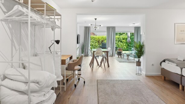 A modern, well-lit room with a bed, a desk, and a chair. There is a window with curtains, a plant, and a view of greenery outside. - Modern KüstenHygge 2 with garden & parking space (Westerdeichstrich)